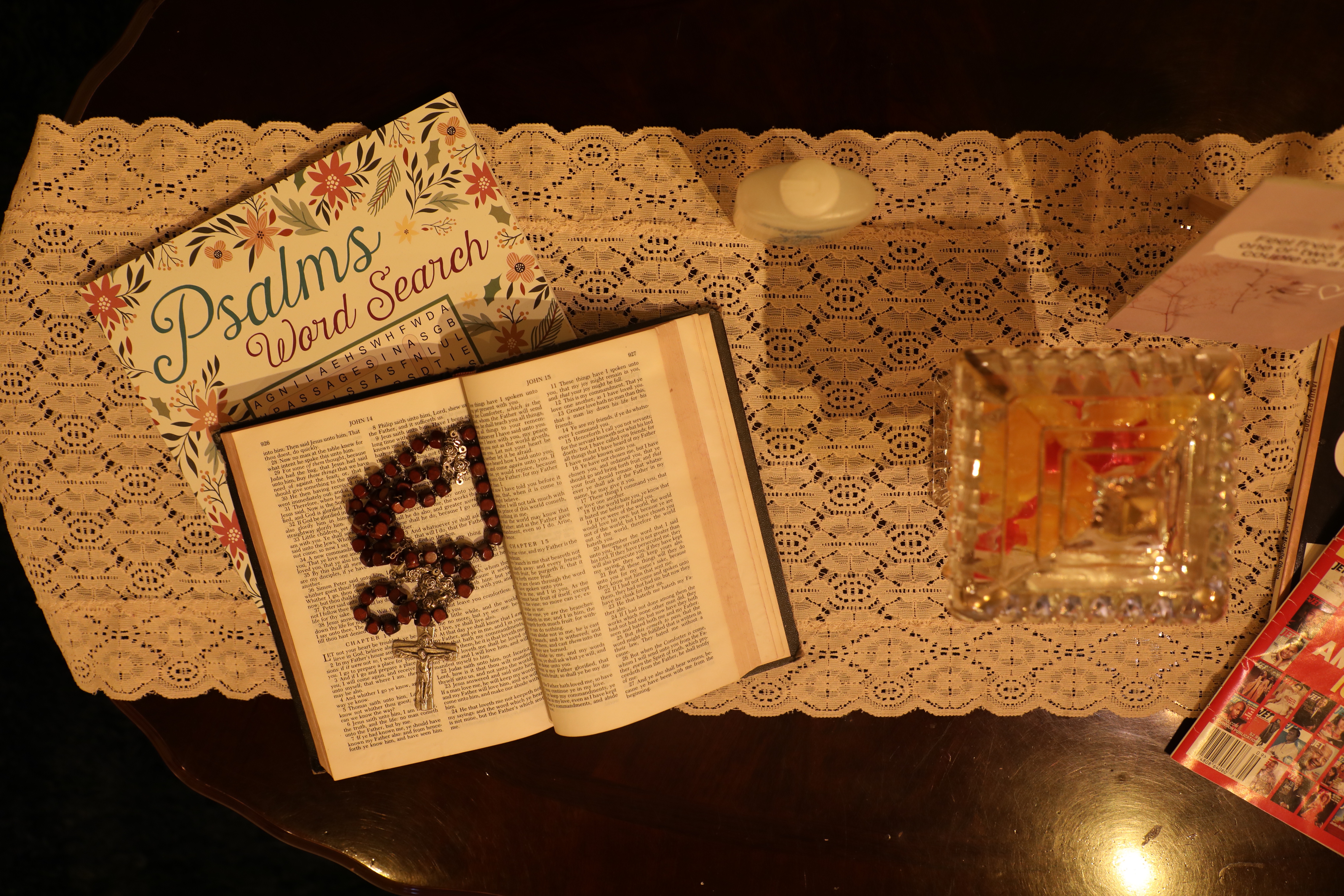 An open Bible with a rosary necklace between the pages. It's sitting on a lace doily besides a candy dish and Psalms word search. 