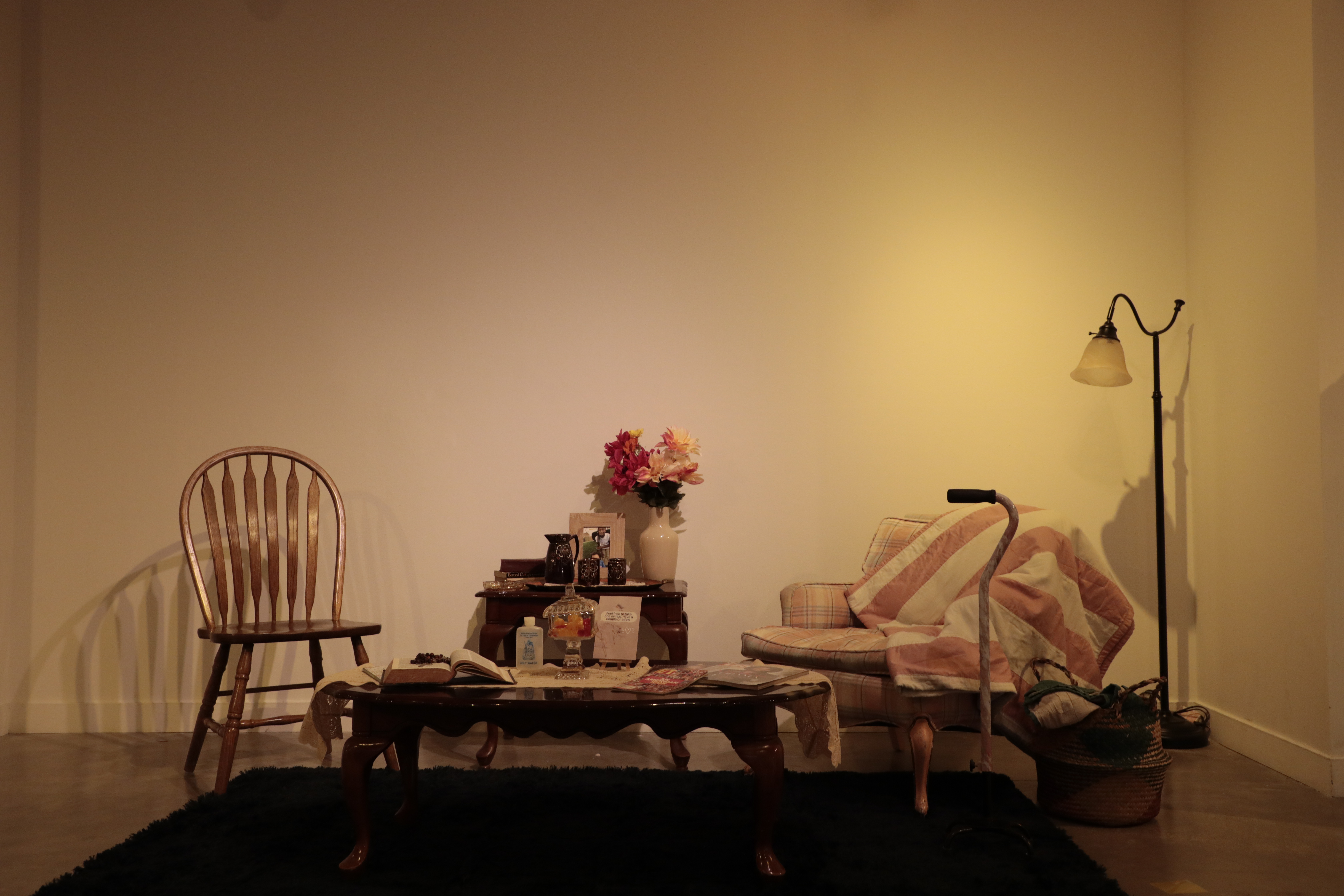 A wooden chair on the left and a cushioned arm chair with a blanket wrapped over on the right. Two wooden tables sit in the middle holding an open Bible, a vase of flowers, and family photohgraphs. 