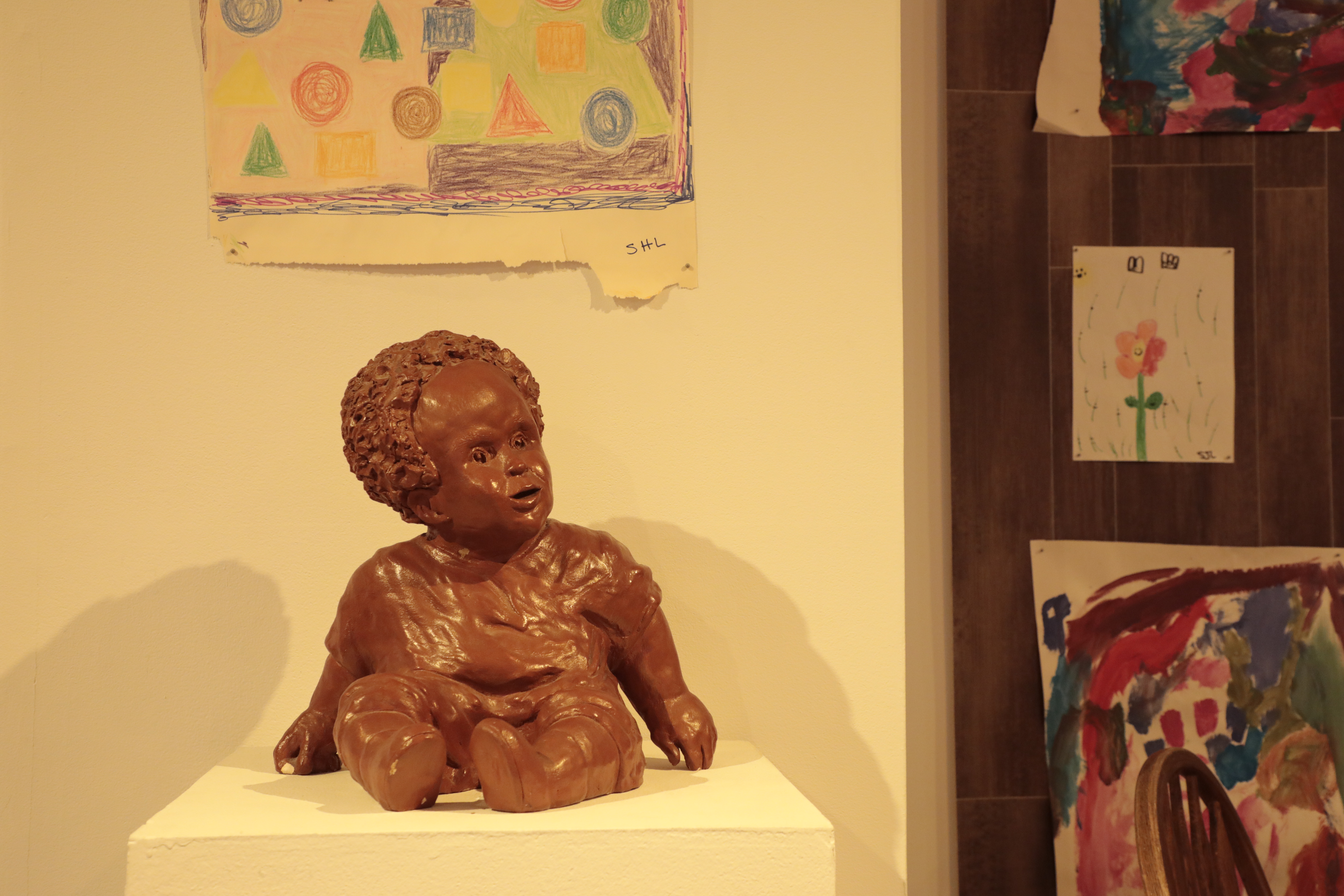 A ceramic statue of a baby with children's drawings on the wall behind the statue. 