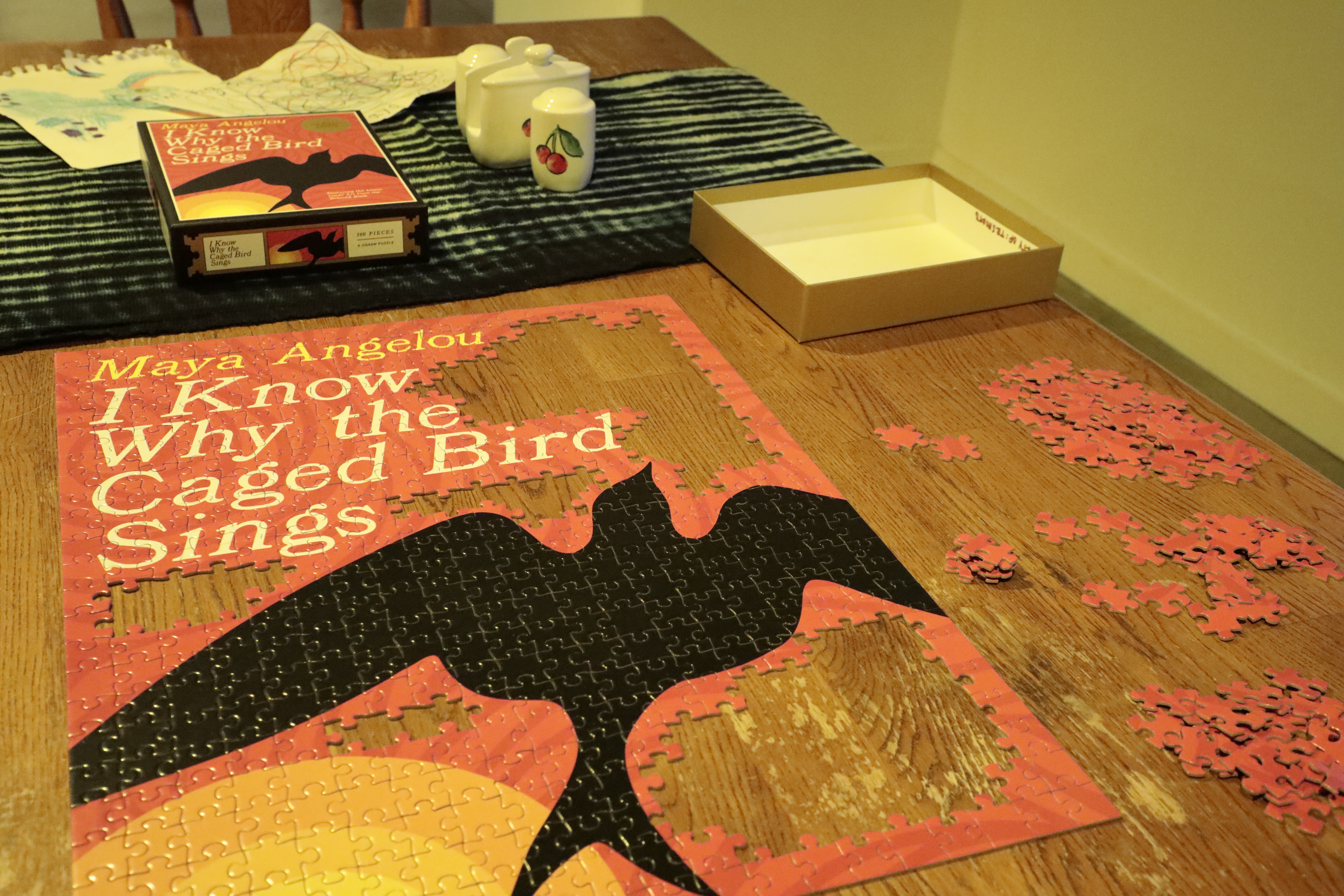 A half complete puzzle with pieces scattered across the table. The puzzle depicts a bird and sun with the the words "Maya Angelou: I Know Why the Caged Bird Sings" 
