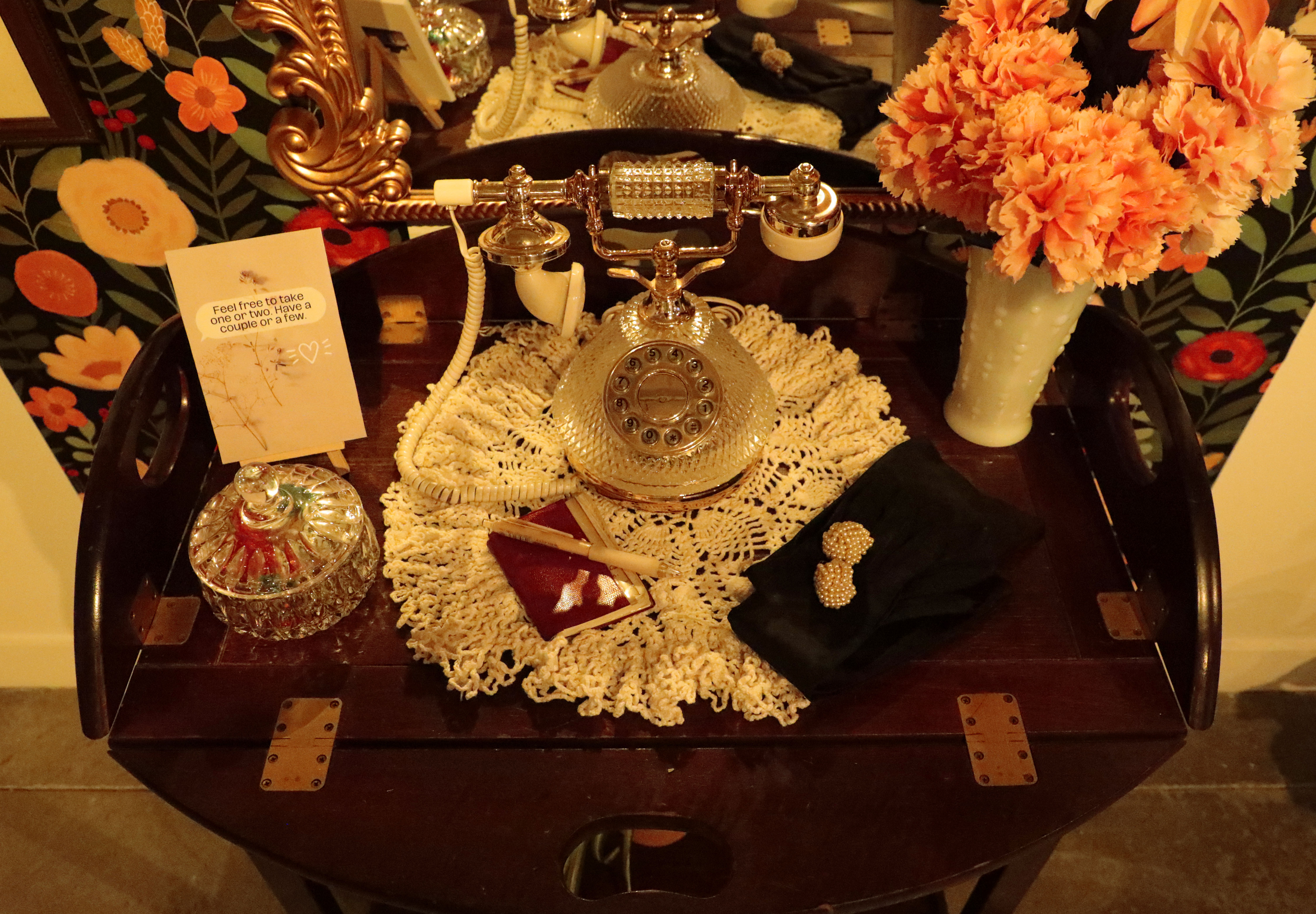 Gold rotary phone sitting on doily on a table surrounded by a jar of old fashioned candies and a vase with orange flowers, all lit by warm tones