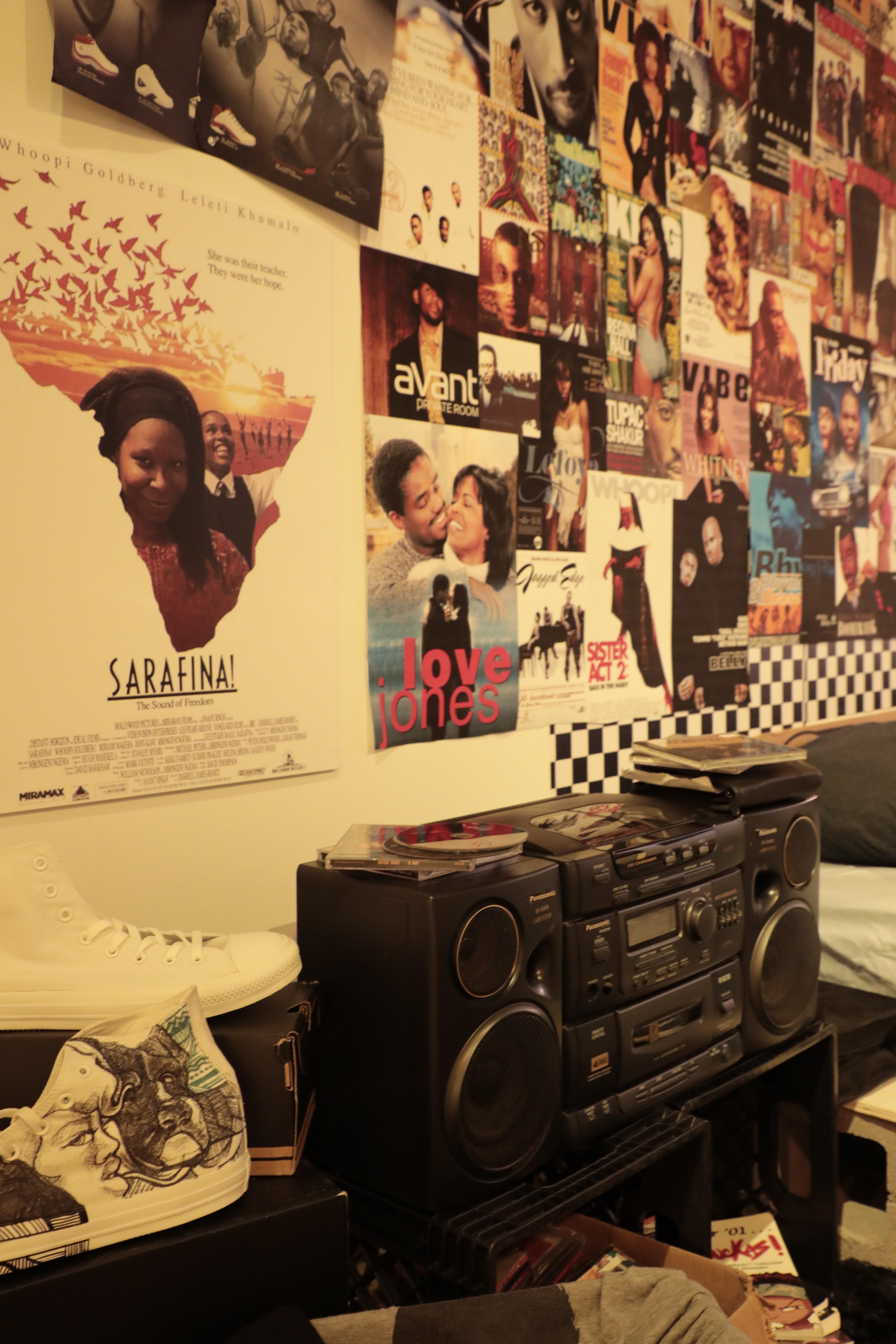 A stereo with CD's stacked on top. Shoes sit beside it with a large collage wall in the background. 