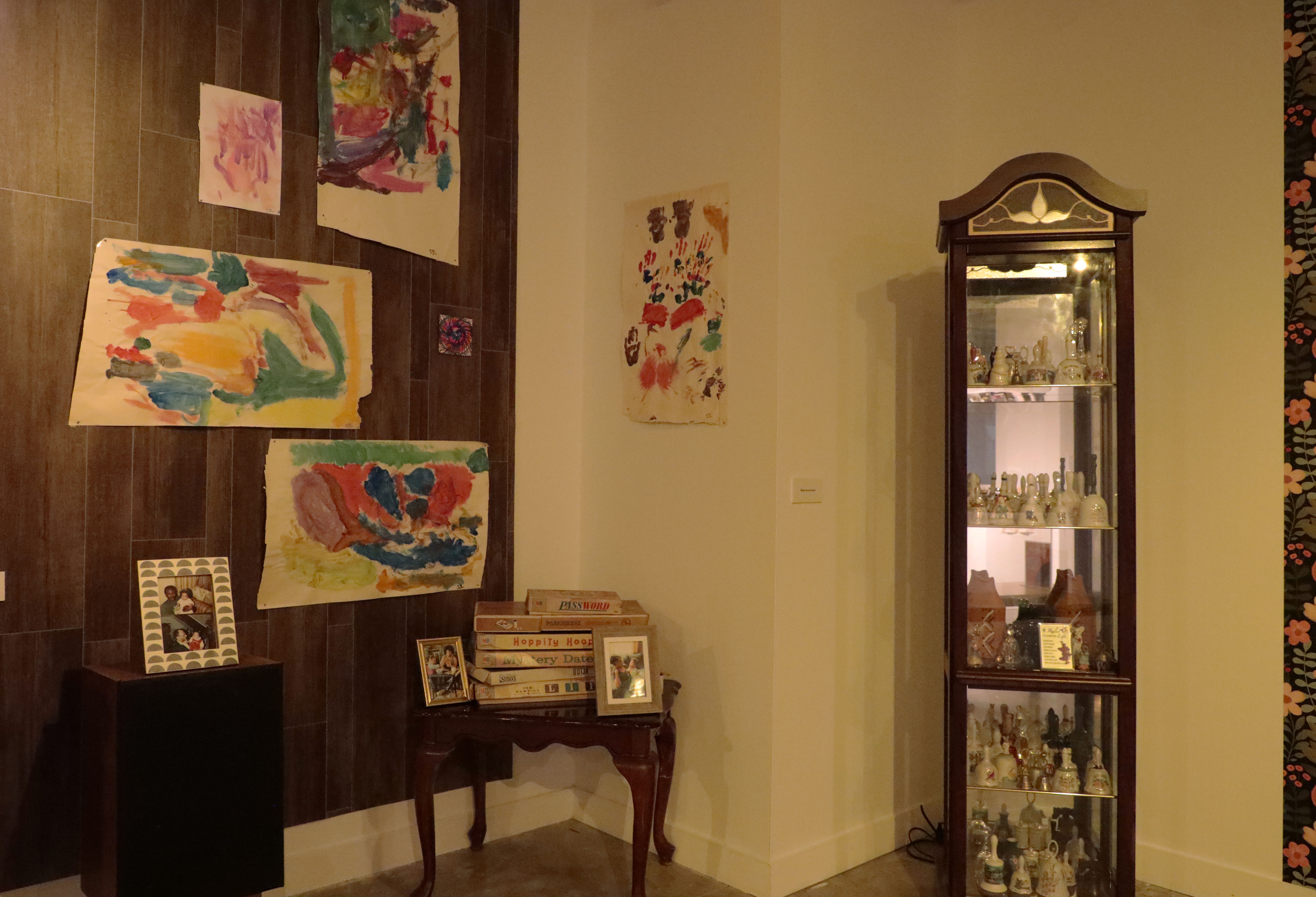 A glass cabinet on the right side full of glass bells. Children's painting is hung on the wooden paneled wall to the left 