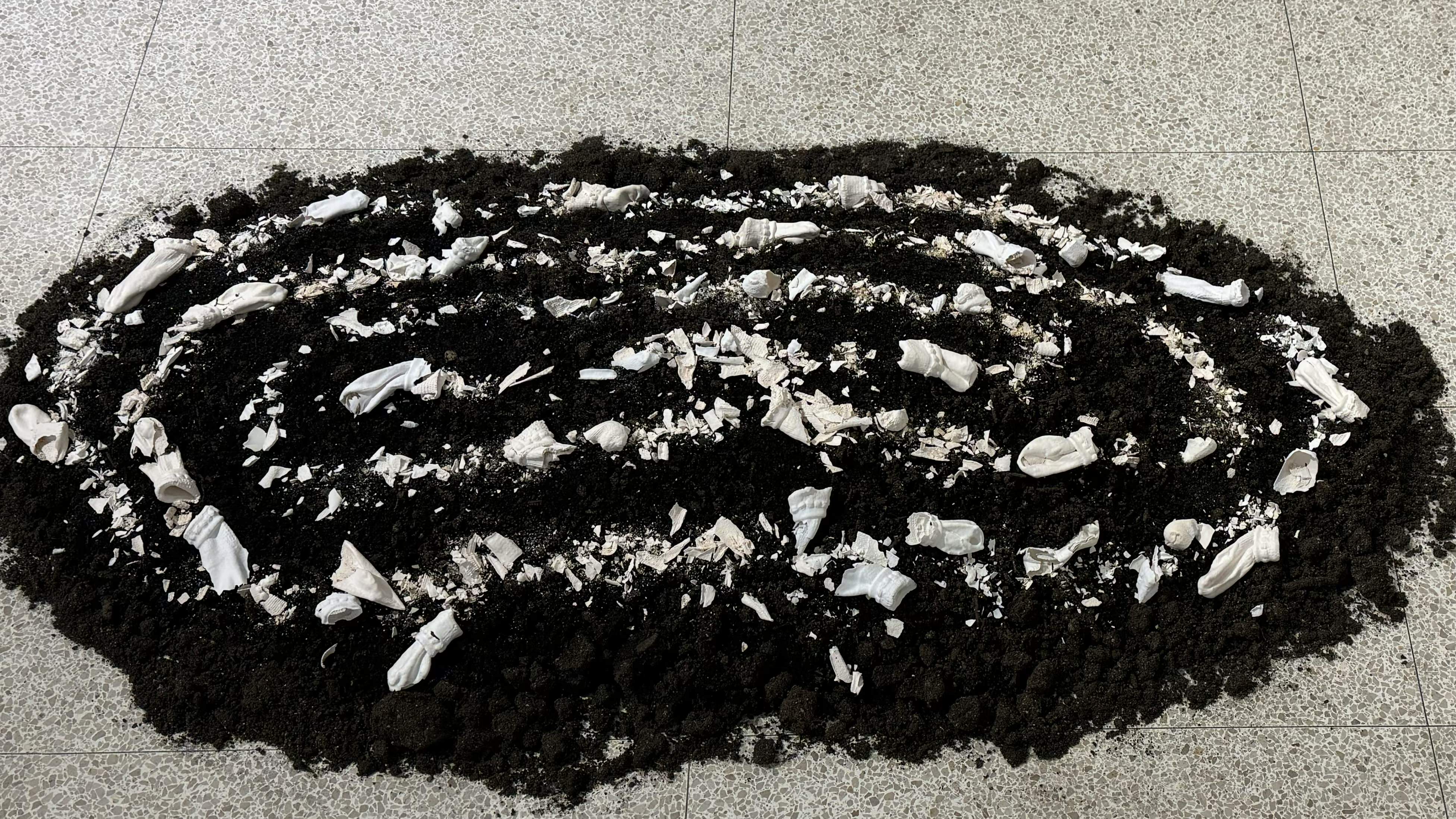 A top-down wide shot of the art installation, showing the oval mound of dark soil contrast against the light floor, scattered with delicate white ceramic pieces that resemble folded cloth and broken pottery.