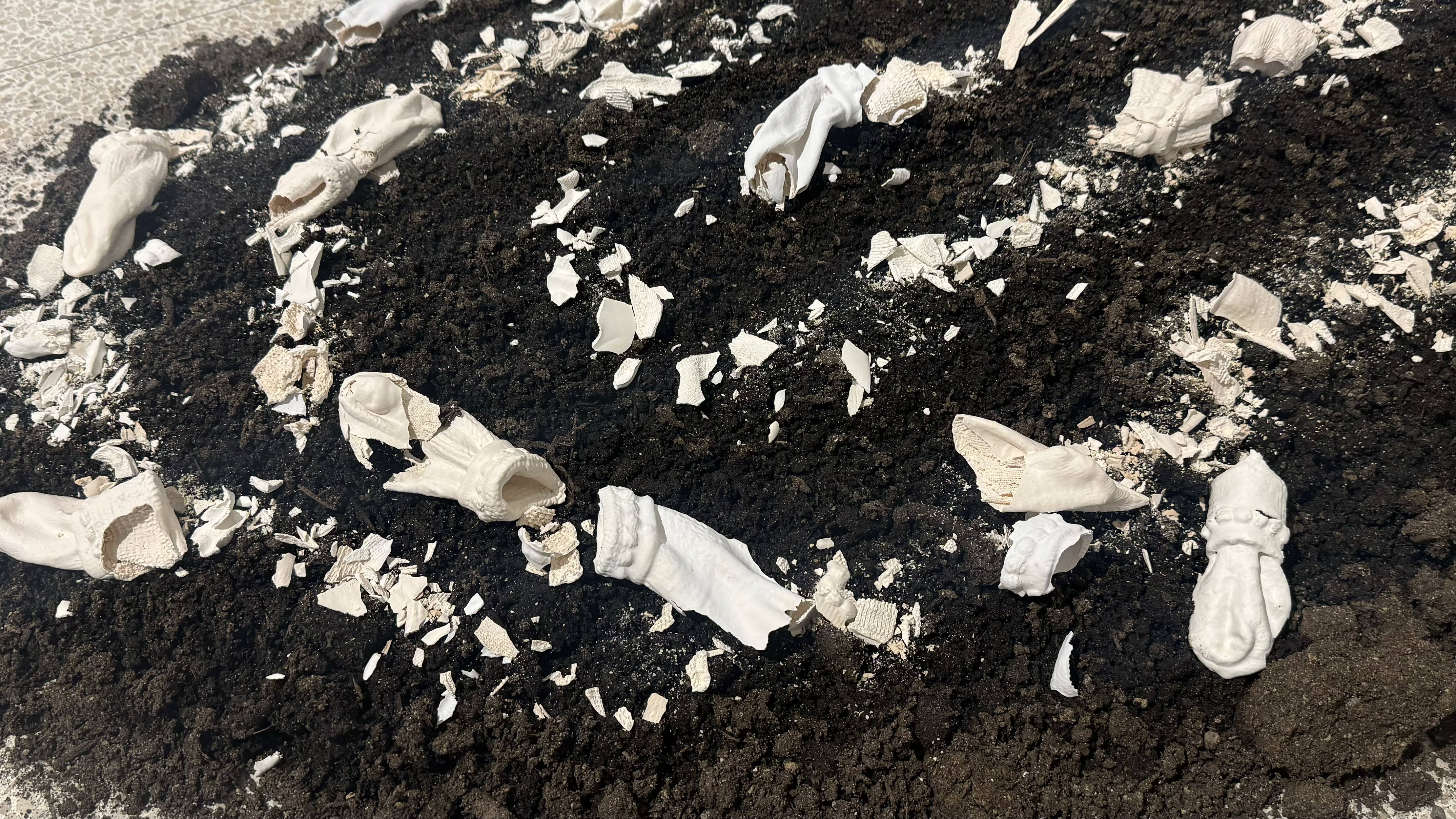 An installation art piece featuring broken white ceramic or plaster fragments and cloth-like sculptures scattered across a mound of dark, rich soil.