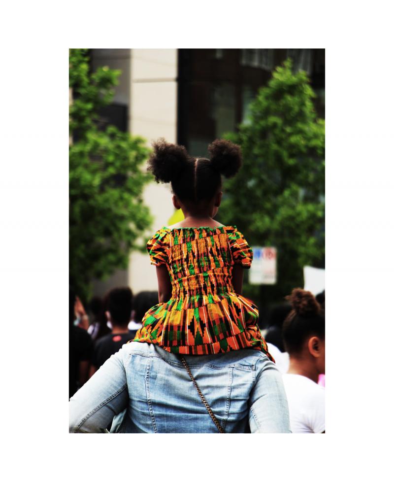 Young Black girl sitting on a person's shoulders in a brightly patterned dress.