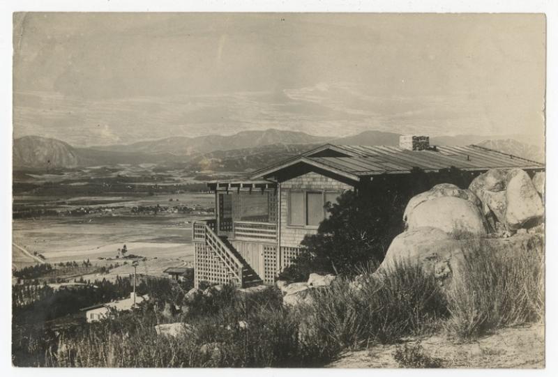 A black-and-white photograph of a house resting on a hill in front of a large field