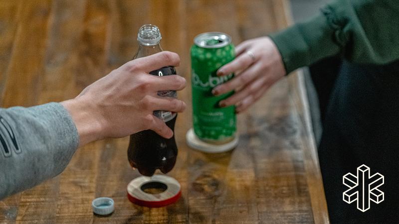 A hand holds a bottle of soda over a small wooden circle with ah ole in the middle, and one hand holds a can of sparkling water on another coaster.