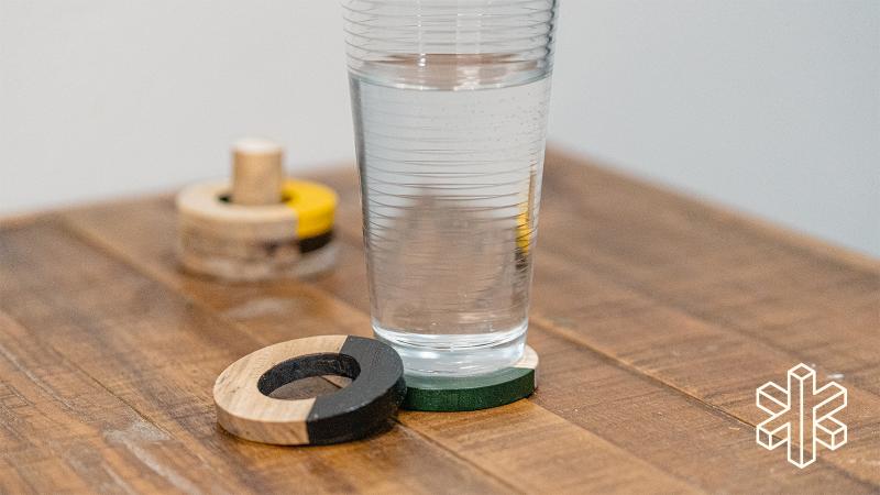 A glass of water sits on a table next to a small wooden circle with a hole in the middle that is half painted black. A few more circles are stacked on a wooden peg in the background.