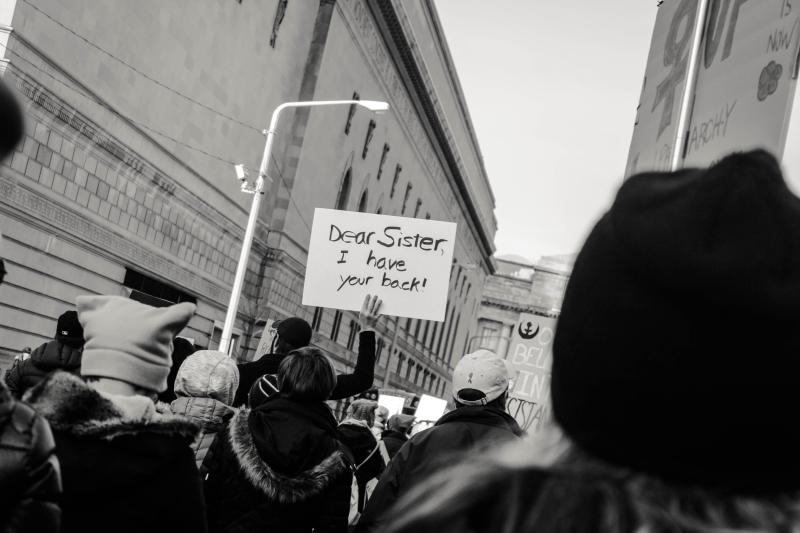 Group of people protesting on street in winter. One sign says, “Dear Sister, I have your back.!”