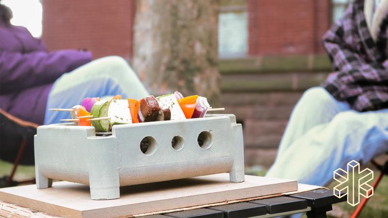 A small concrete grill sits on an outdoor table with kabobs stacked with food slices on top.