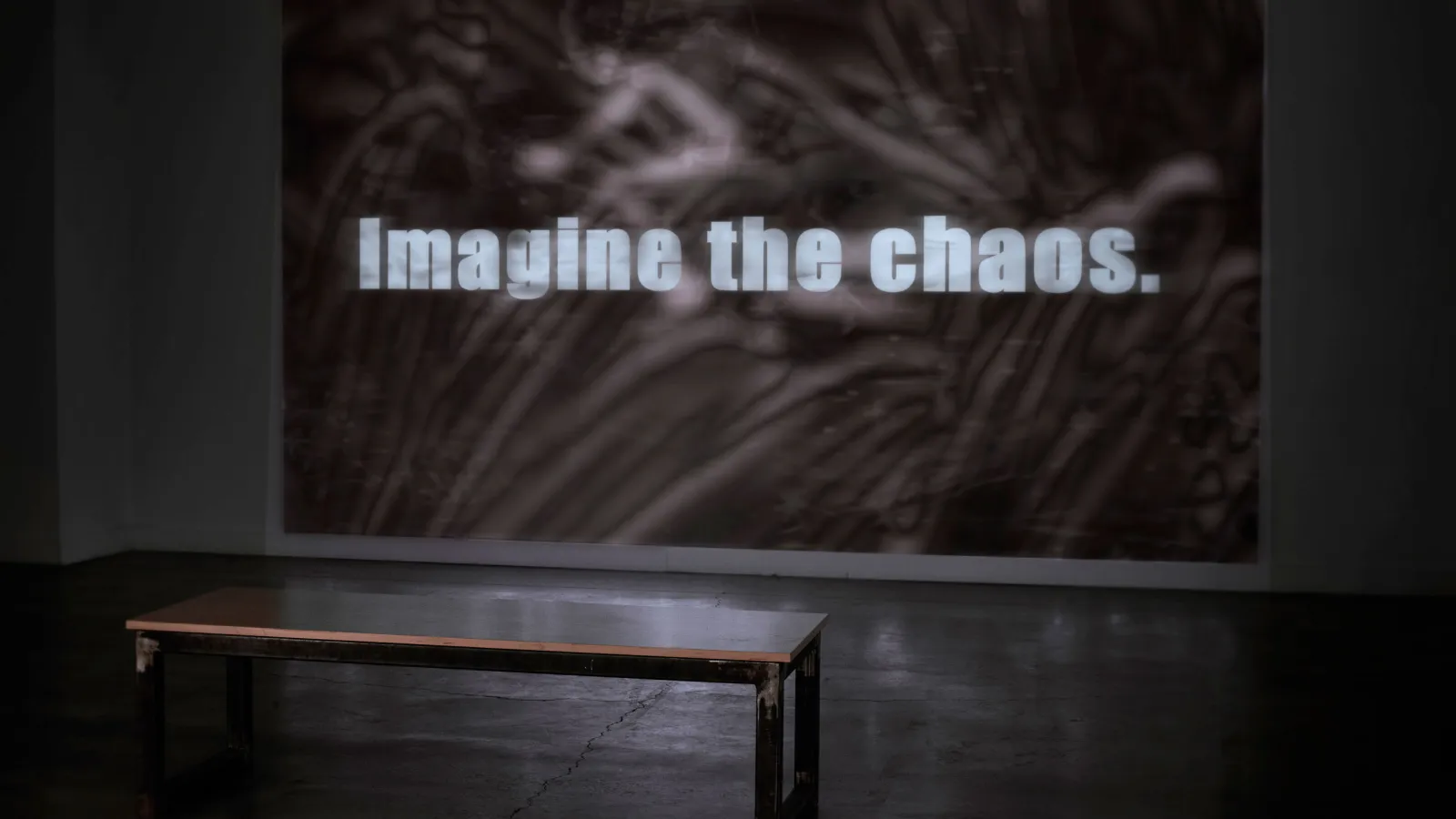 a wall with a video projected onto it with a bench nearby, projected video still says "imagine the chaos"