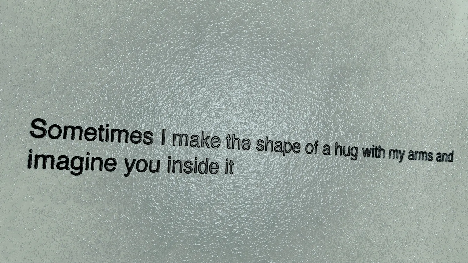 Black text on a bumpy grey background reads "Sometimes I make the shape of a hug with my arms and imagine you inside it"