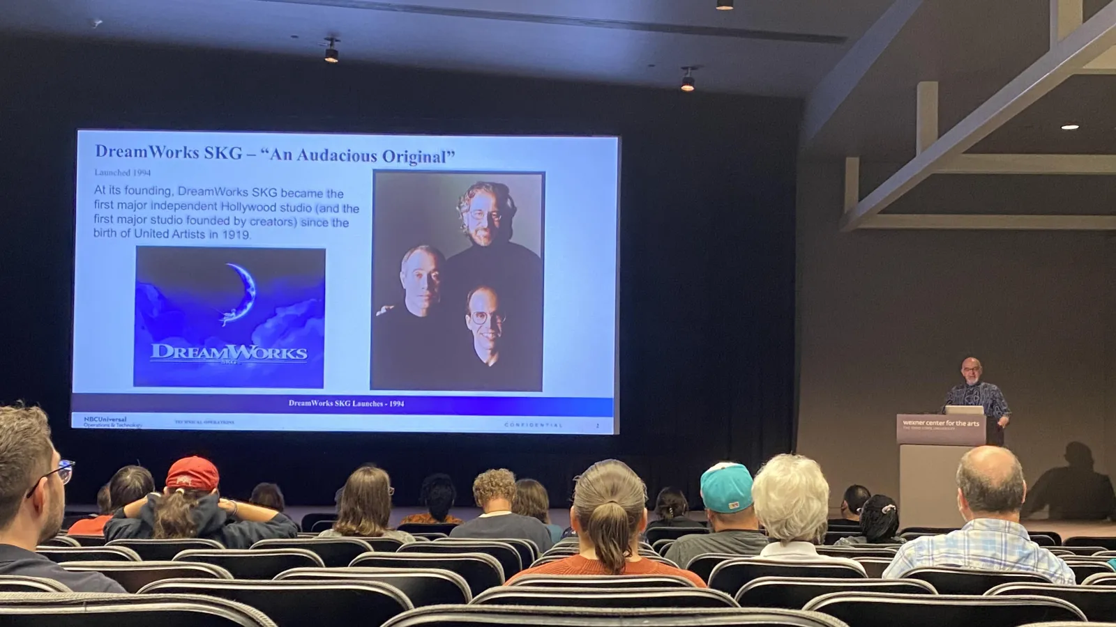Facing a stage towards the back of an auditorium, Chase Schulte stands at a podium on the right facing the audience, with a large PowerPoint slide projected in the middle of the room. The slide shows a group picture of Steven Spielberg, Jeffrey Katzenberg, and David Geffen on the right, with the title "DreamWorks SKG – "An Audacious Original"" on the upper left. Below this is the line "Launched 1994" and a body text that reads "At its founding, DreamWorks SKG became the first major independent Hollywood stu