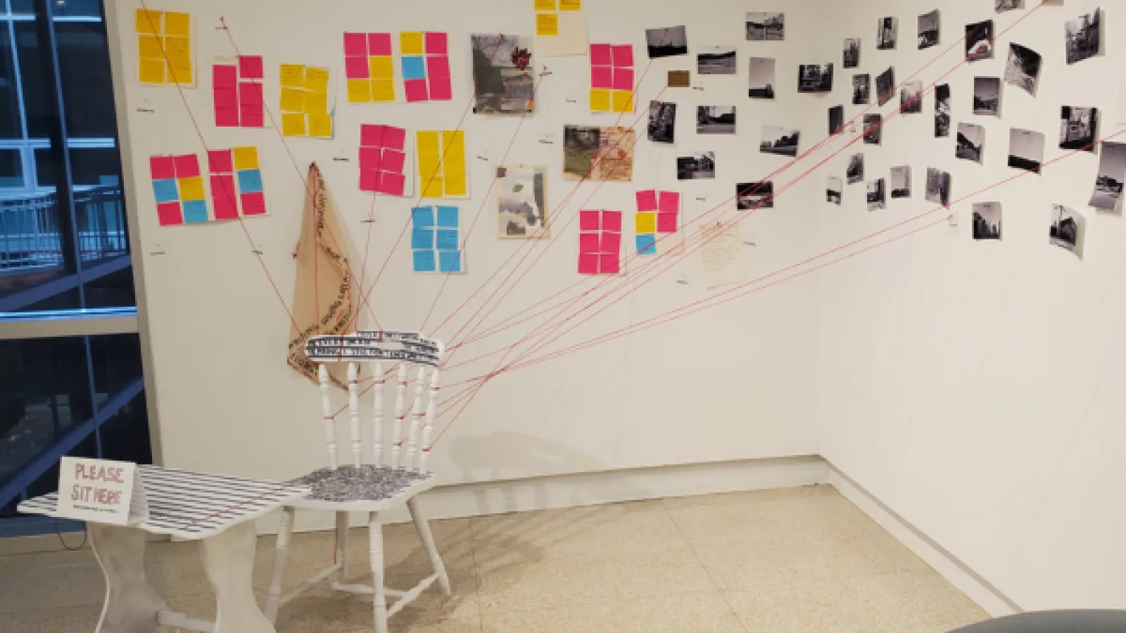 A white chair sits facing away from the corner of the gallery. Red strings attached to the back of the chair are connected to the wall. On the wall are multicolored sticky notes and black-and-white photos.