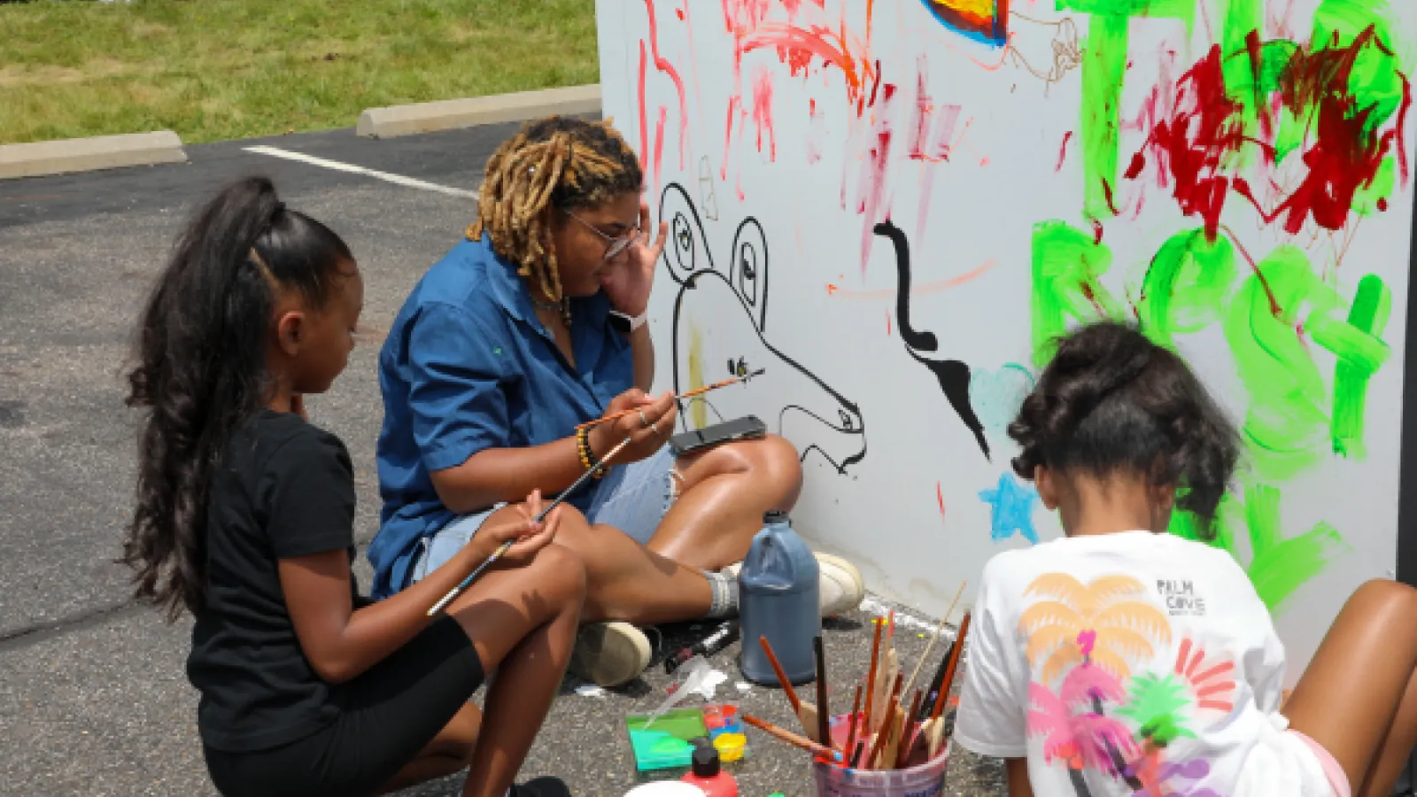 An adult and two children sitting on the ground paint a mural on a white wall together.