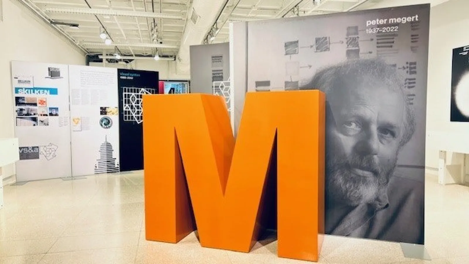 Peter Megert exhibition with panels including one of Peter along with a large orange letter M