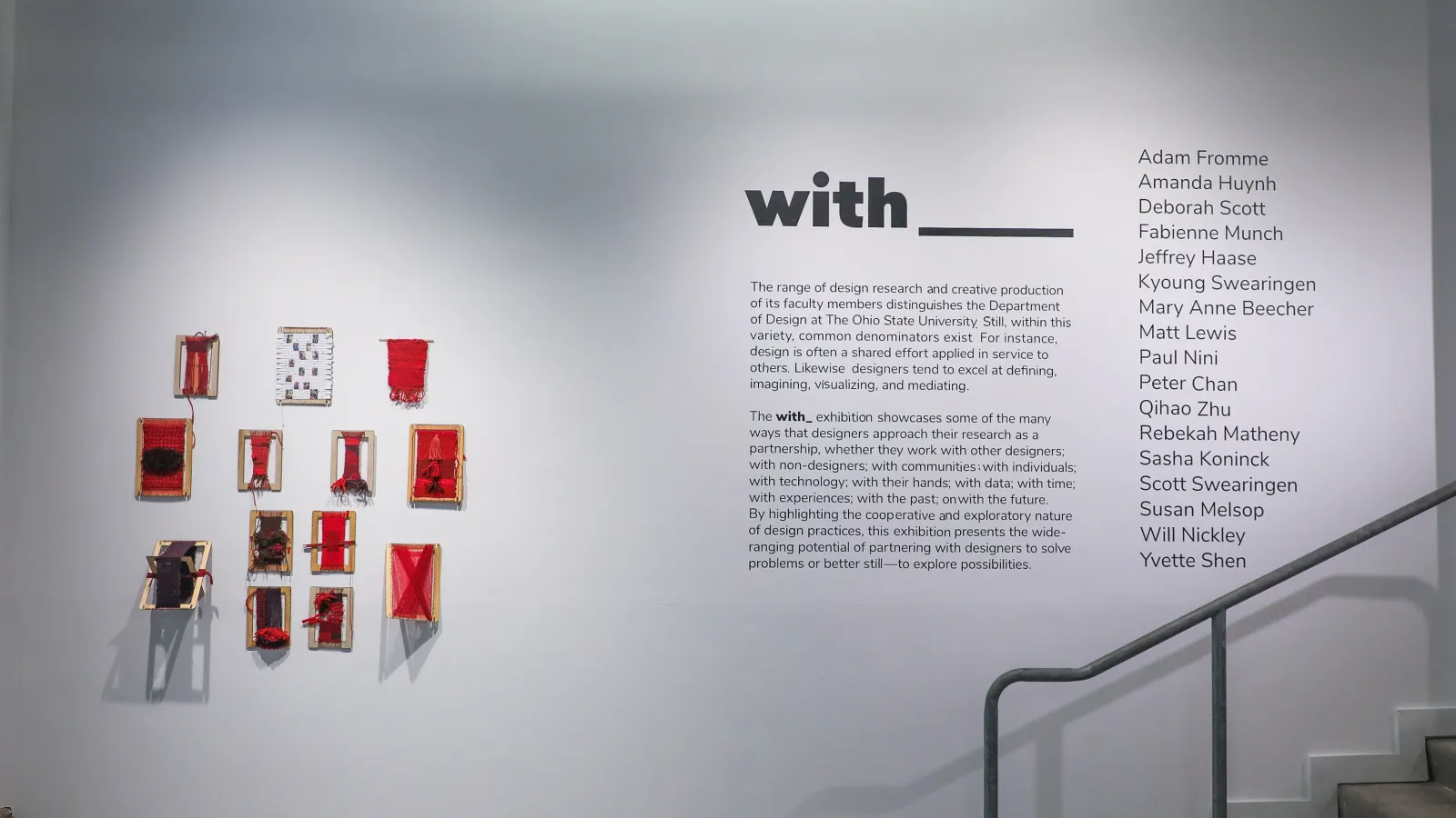 Thirteen rectangle weaving looms are spread over the wall. The yarn weaved through the looms are various shades of red. The weaving looms are next to the exhibition's vinyl description.  