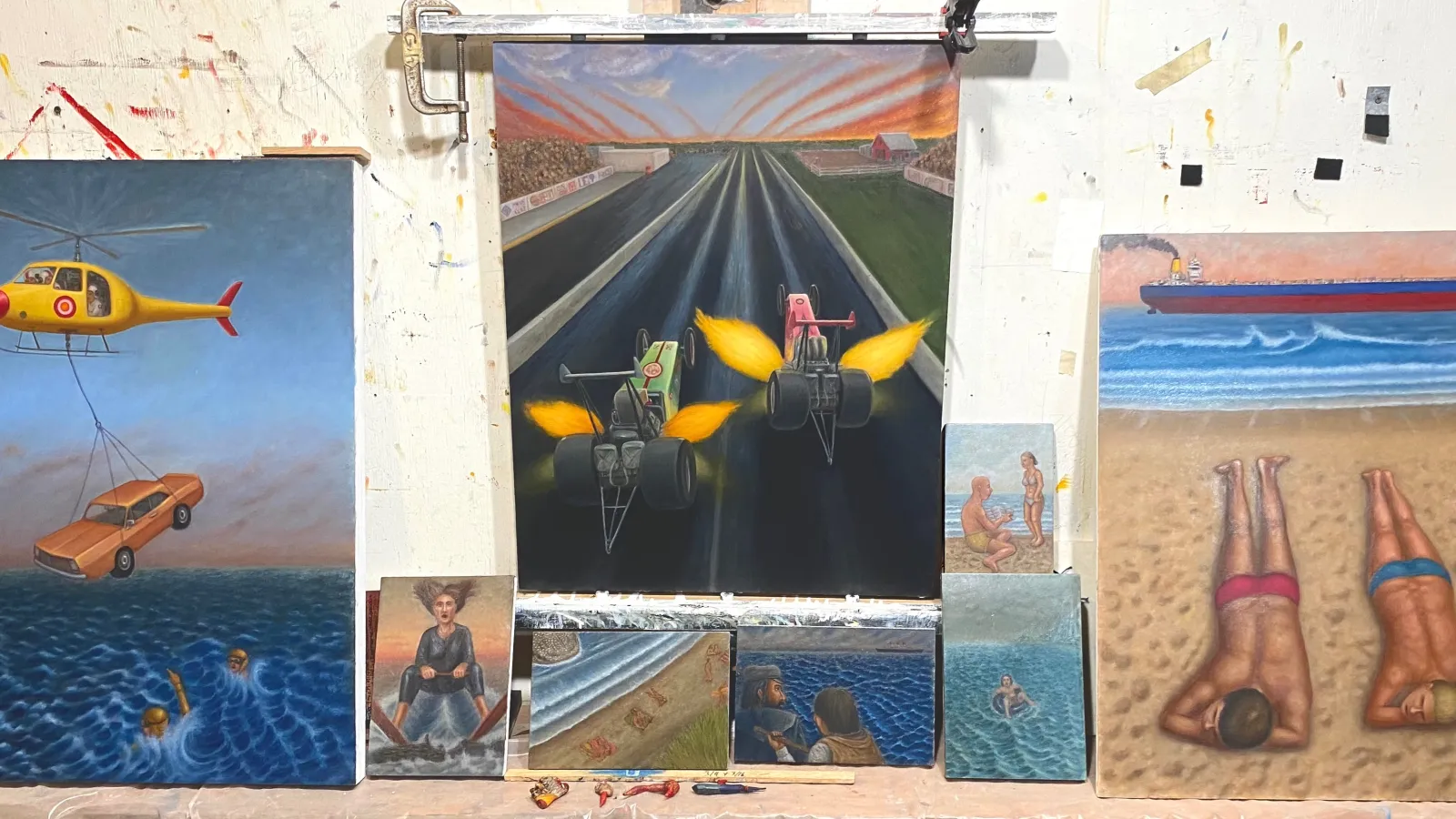 three paintings by glen holland, including one of a helicopter lifting a car; one of two race cars; and one of two people sunbathing on a beach