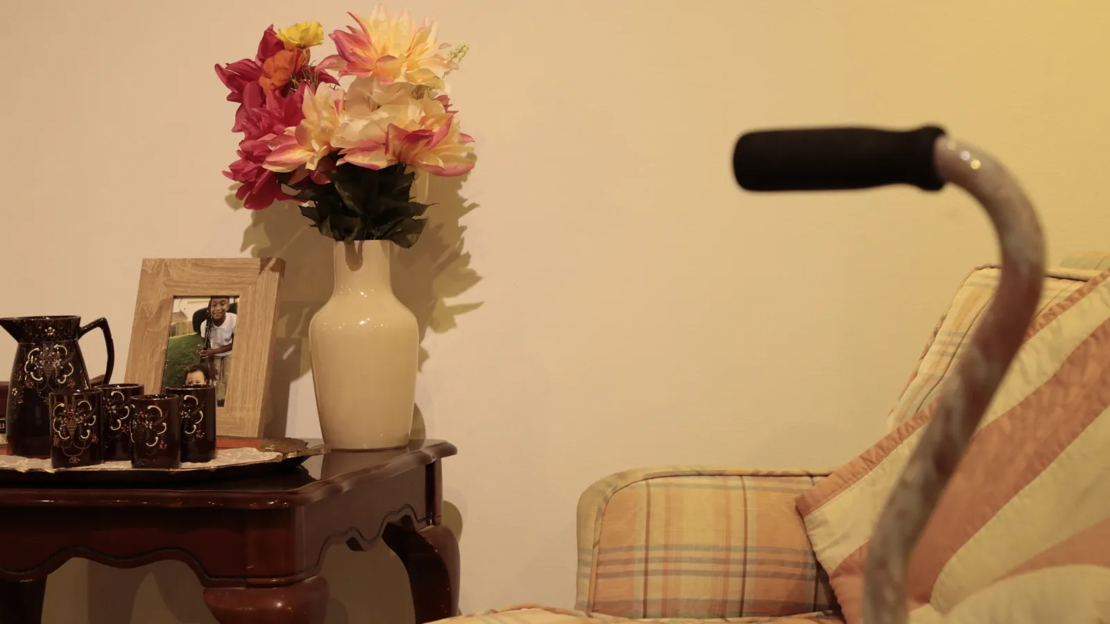 A table with flowers, cups and a pitcher, and a picture frame with an image of two kids. Besides it is an armchair and a cane. 