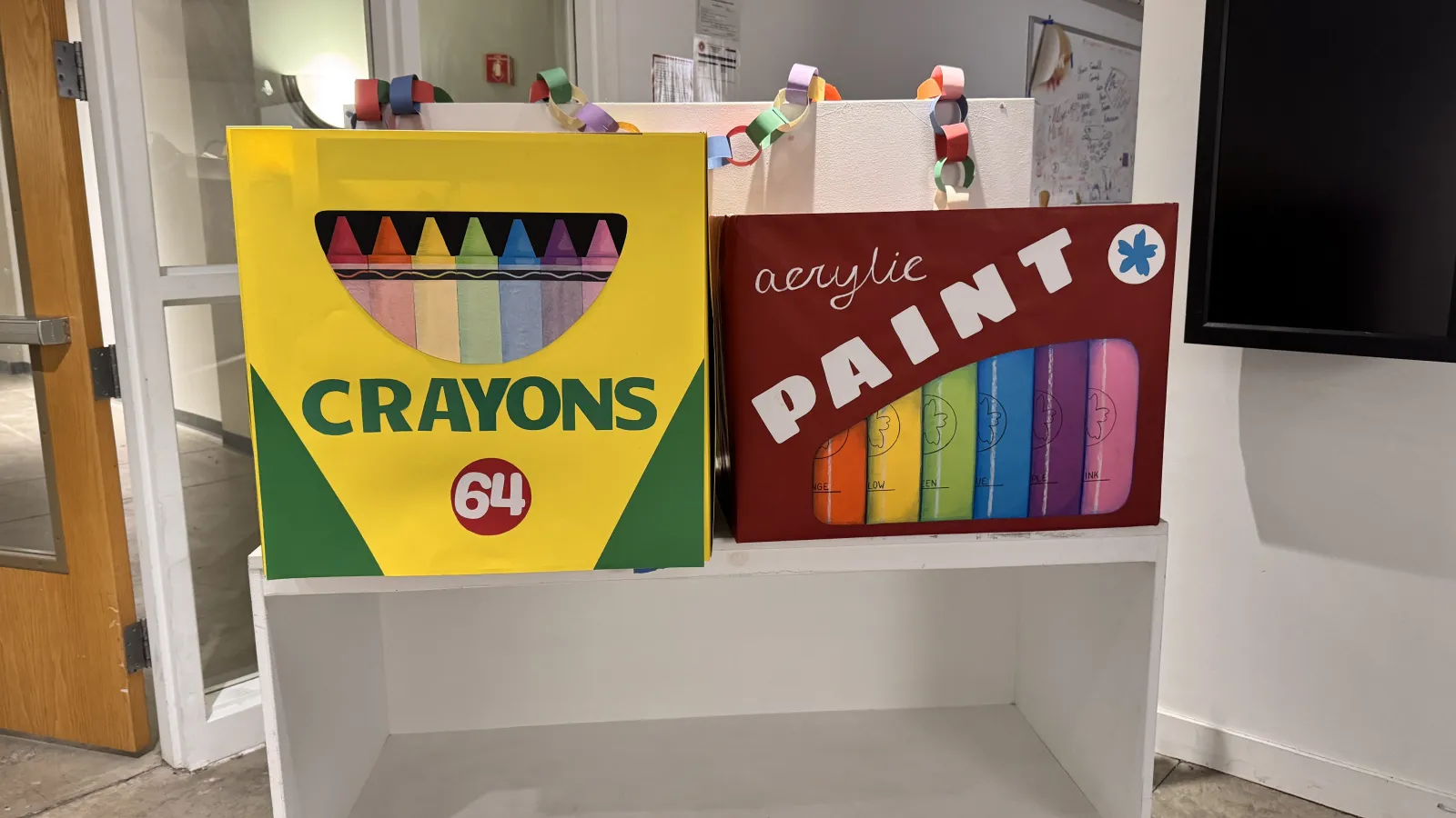 Two oversized, handmade supply boxes—a yellow "Crayons 64" box and a red "Acrylic Paint" box—sitting on a white rolling cart in a gallery.