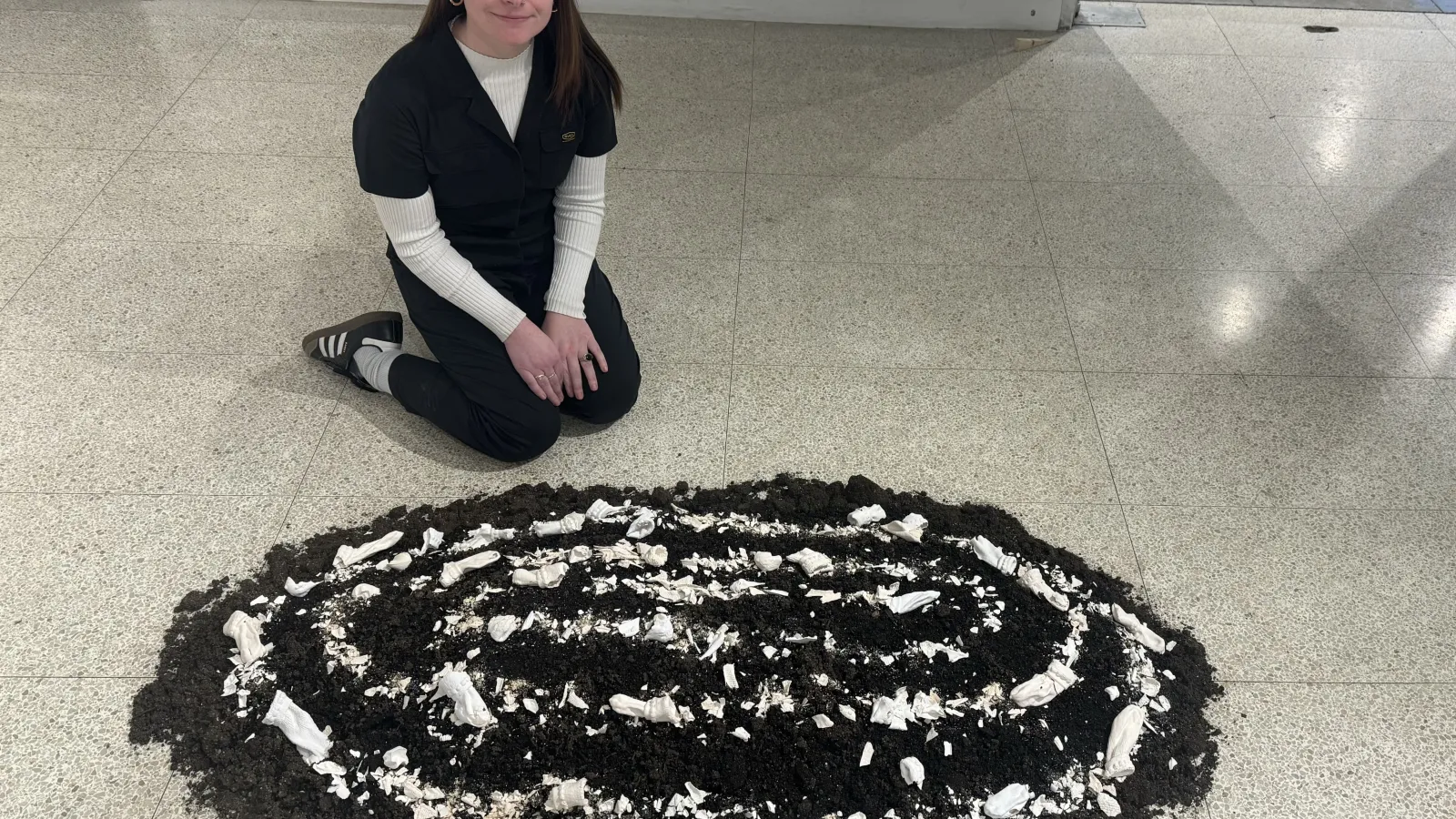 The artist kneels on the gallery floor behind the oval-shaped soil installation, which features concentric rings of white ceramic shards and fabric-like sculptures.