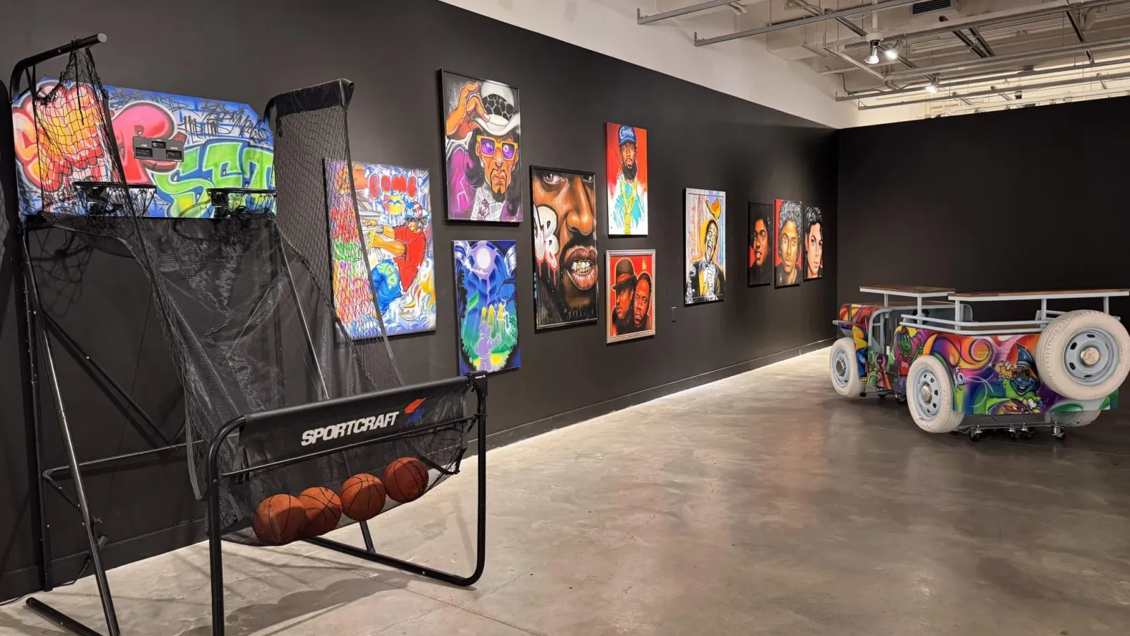 Paintings hung on a black wall with two 3D pieces in front.