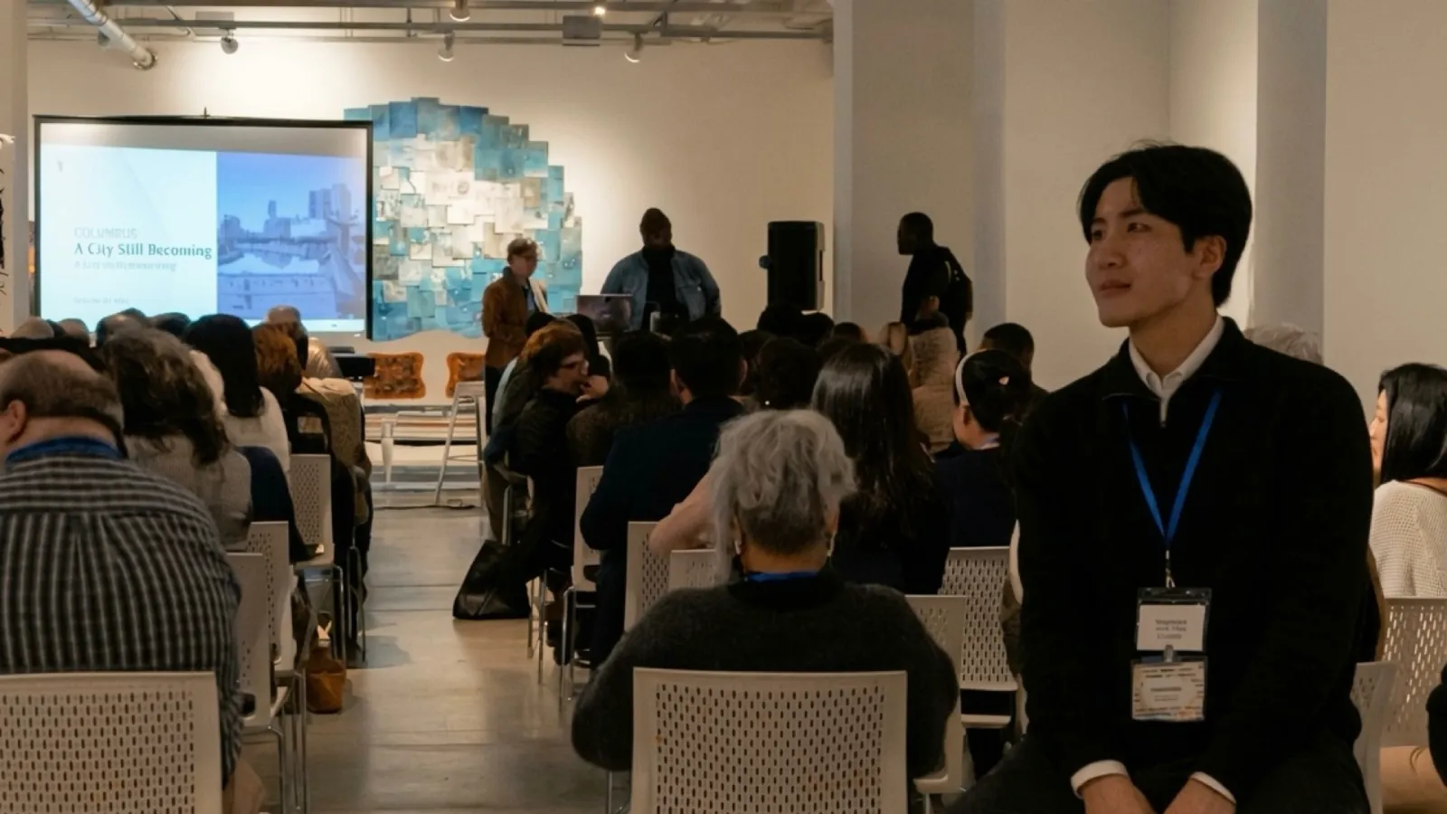 An audience seated in an art gallery watching a presentation displayed on a projection screen, with Ukheon at the front.