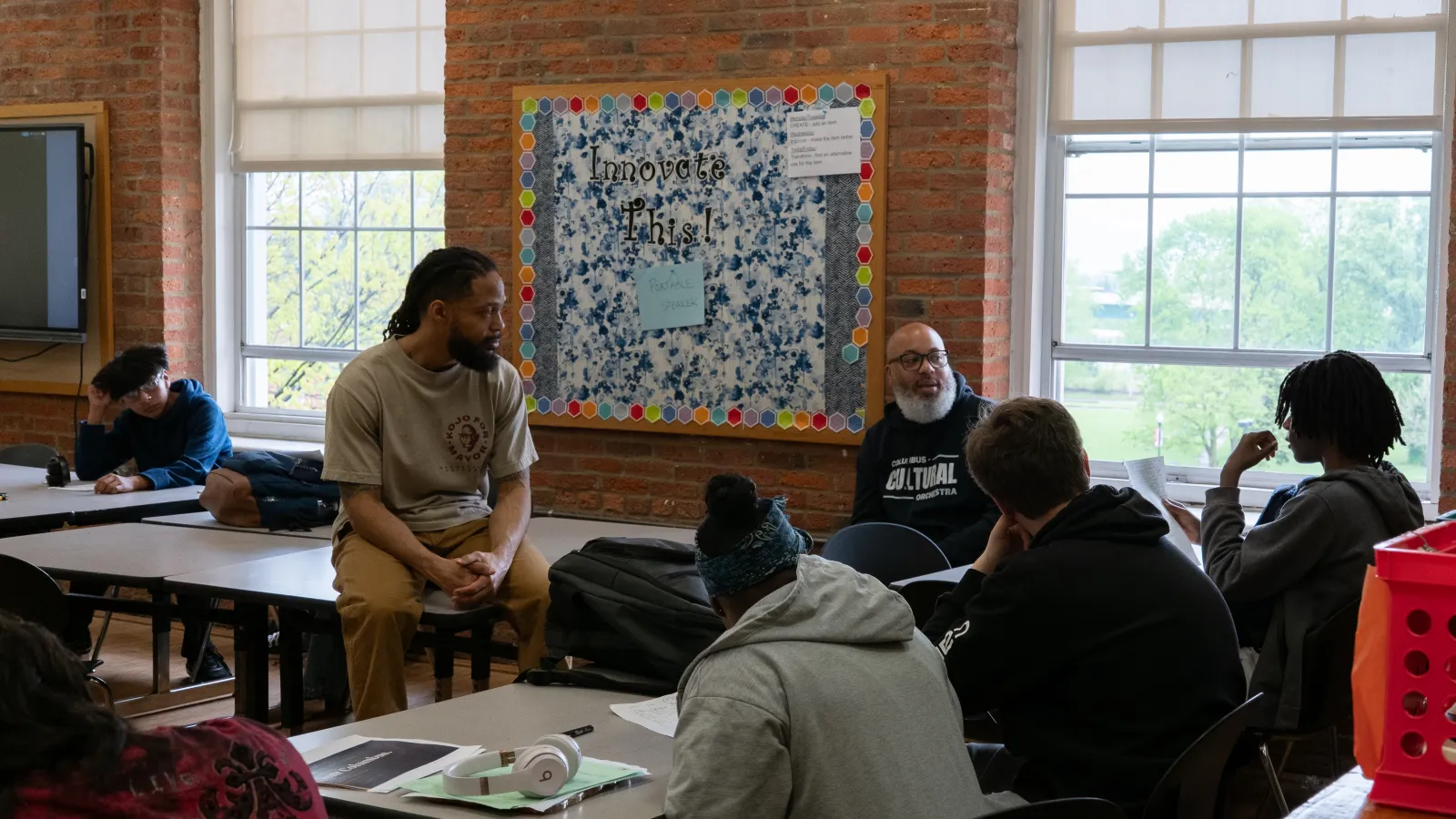 Two instructors sit and talk with students gathered around tables in a classroom.