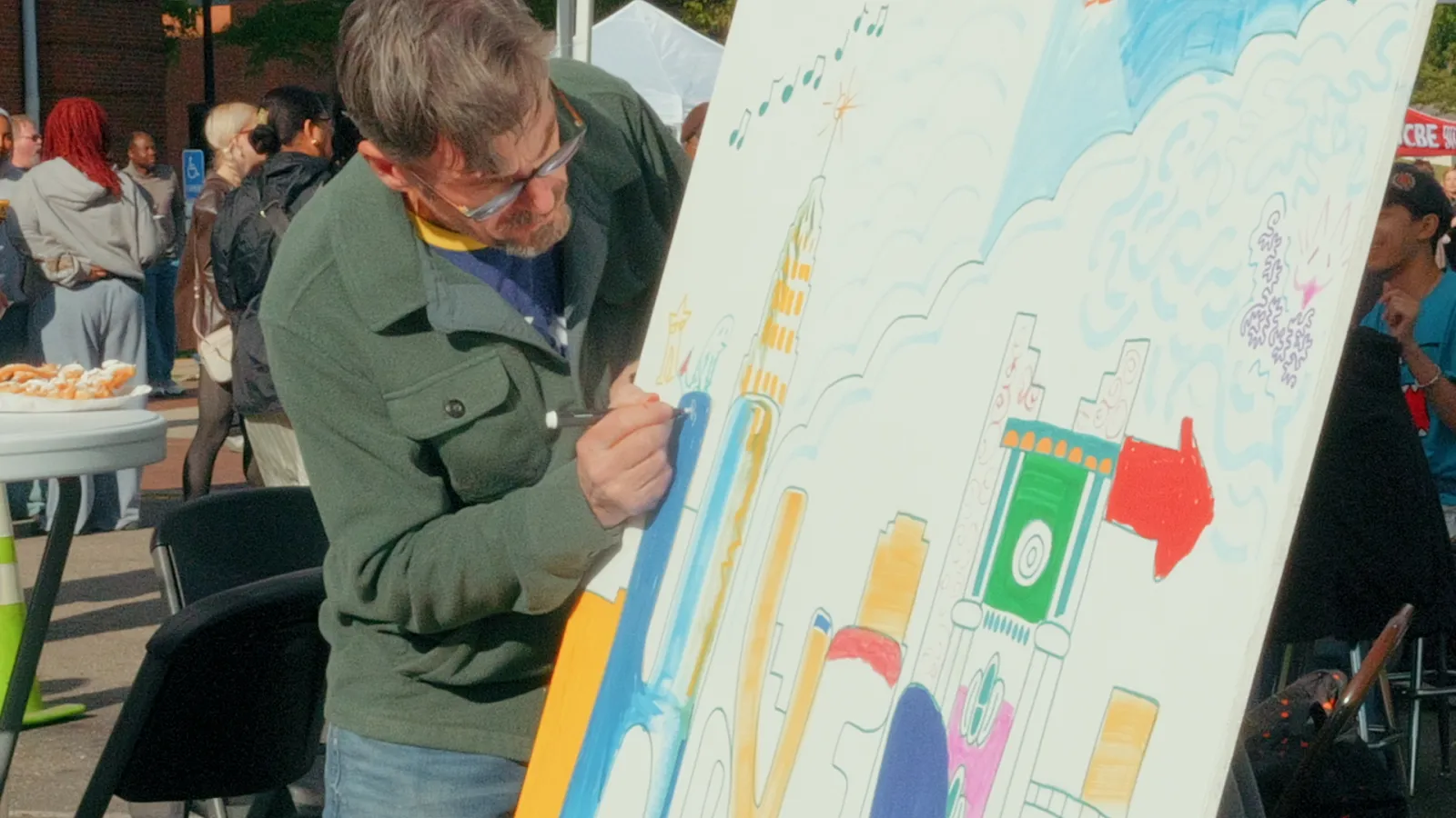 An artist with glasses draws a cityscape illustration on a large white display board outdoors.
