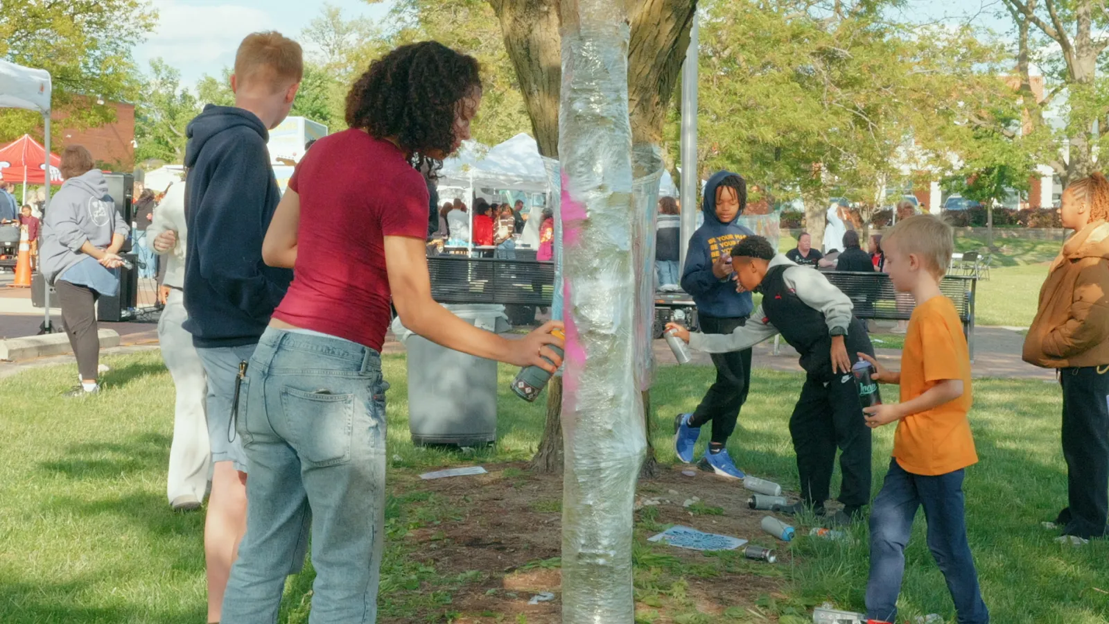 People gather outdoors using spray paint to create art on plastic wrapped around a tree trunk.