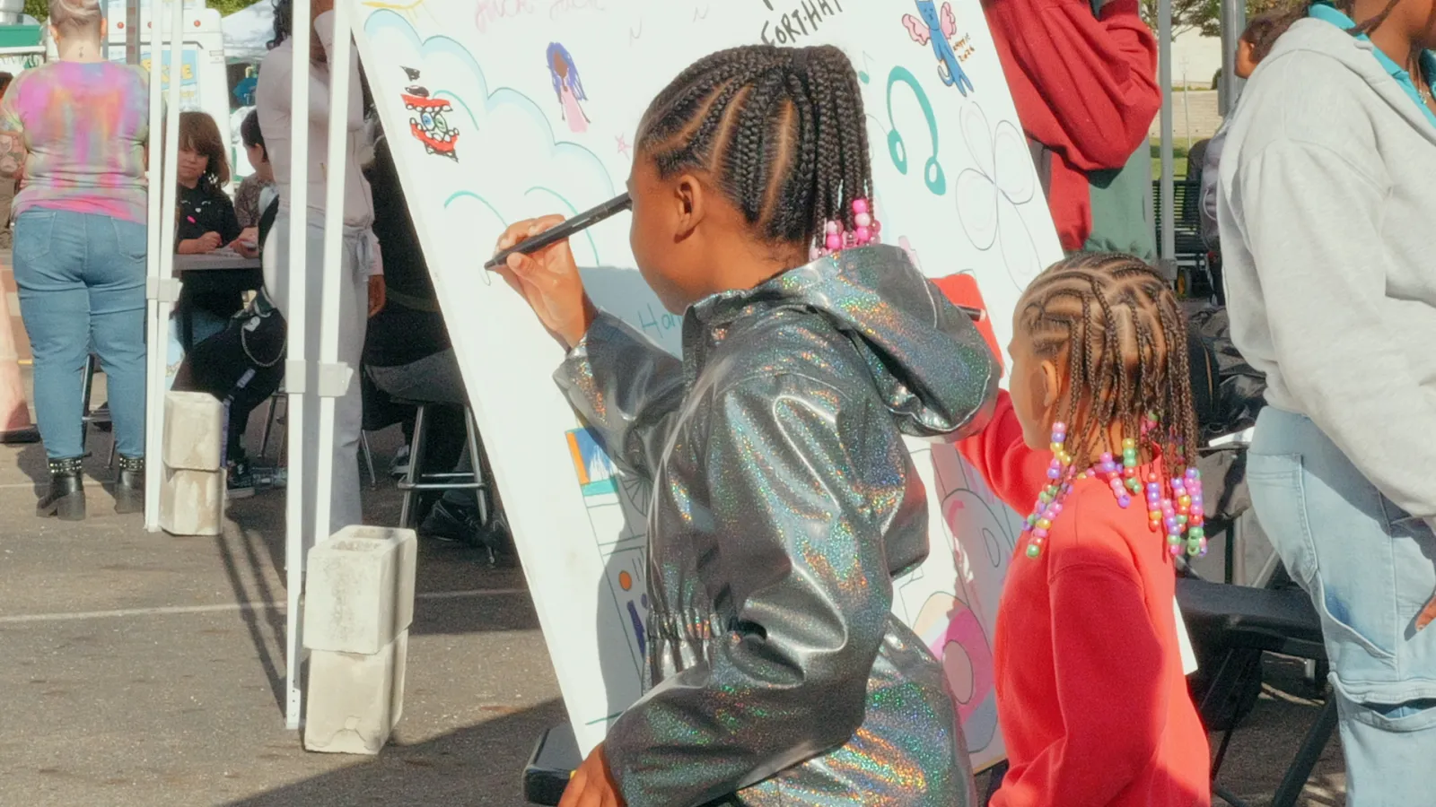 Two young children draw with markers on a large white canvas set up outside.
