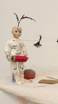 A close-up view of the assemblage. The baby doll covered in black stitches holds a spool of red thread. Its face is cracked and pins are stuck in its body.