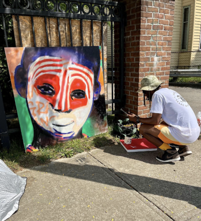 Man on side walk creating a painting of an boy of color with his face painted white and red