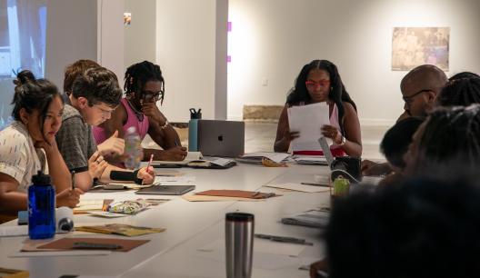 Ajanae at the head of a table teaching a poetry workshop to people