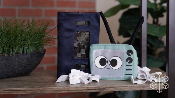 A pouch designed to look like a TV with a cartoon face sits on a table with another black pouch that looks like a VHS tape and crumpled slips of paper.