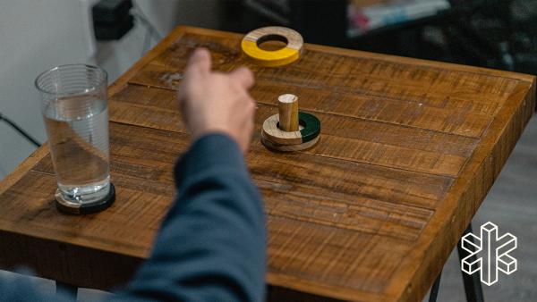 A hand throws a small wooden circle half painted yellow onto a wooden peg sitting on a table with a similar wooden circle painted black stacked on it.