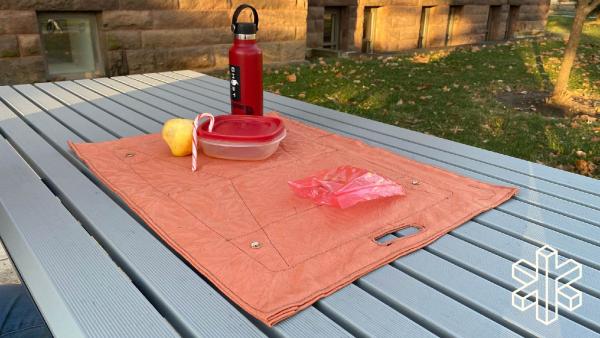The orange mat lays open on a table with a water bottle, apple, candy cane, reusable food container, and plastic baggie sitting on top.
