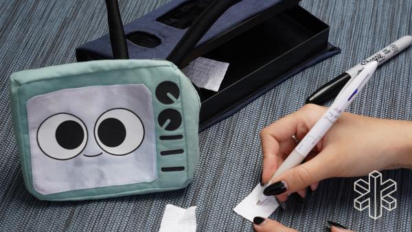 The cartoon TV and black pouch sit on a table. Next to them, a hand uses a pen to write on a slip of paper.