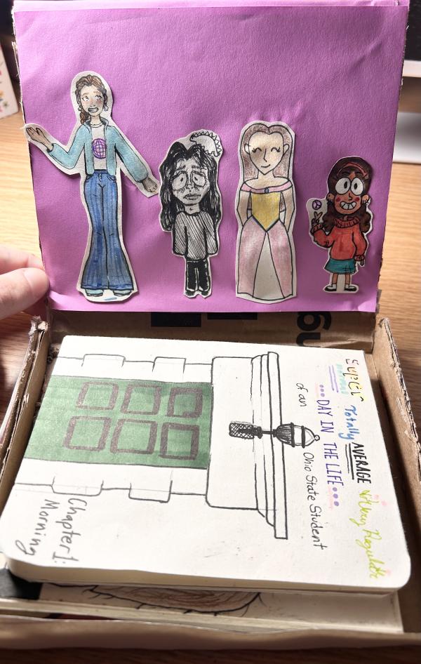 A handmade diorama in a cardboard box featuring a pink backdrop with four hand-drawn, cut-out characters in various styles. Below them, a notebook sits in the box, titled "Super Totally Average Very Regular DAY IN THE LIFE of an Ohio State Student," with a drawing of a building and the words "Chapter 1: Morning."