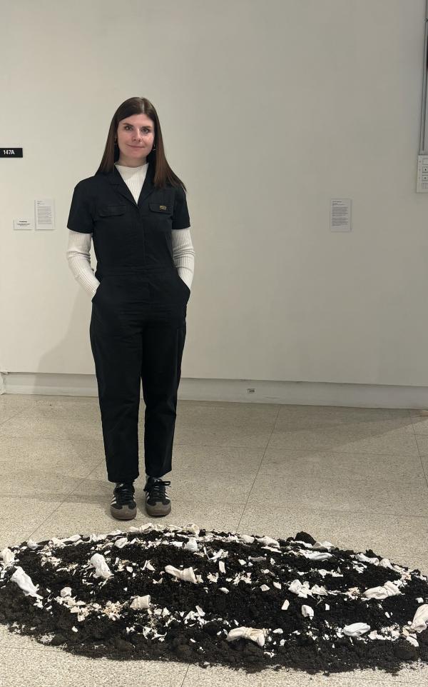 The artist n a black jumpsuit stands behind a circular floor installation made of dark soil and scattered white ceramic shards in a white-walled gallery.