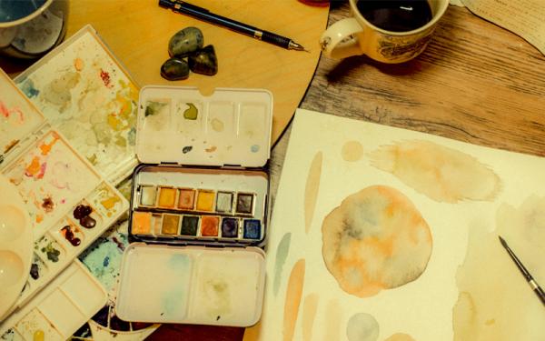 A watercolor palette and painting sit on a desk.