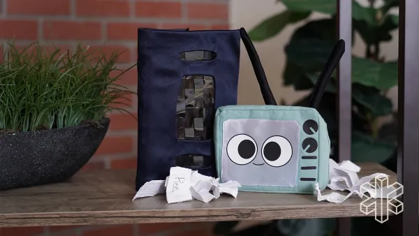 A pouch designed to look like a TV with a cartoon face sits on a table with another black pouch that looks like a VHS tape and crumpled slips of paper.