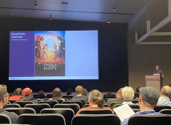 Facing a stage towards the back of an auditorium, Chase Schulte stands at a podium on the right facing the audience, with a large PowerPoint slide projected in the middle of the room. The slide shows a poster for the 1998 DreamWorks film, The Prince of Egypt. On the left of the slide is a label that reads "DreamWorks Animation Theatrical Feature Releases."