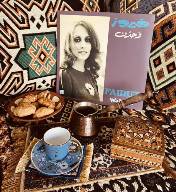 A hand pouring tea next to a dessert and album of Fairuz in front of a backdrop