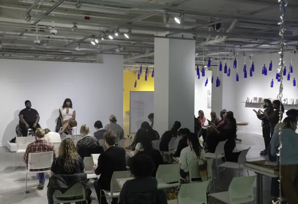 two people sit at the front of the room while a crowd of people in chairs with backs against the camera look to their laps