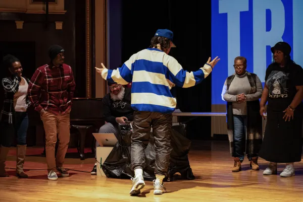 six individuals stand on a wooden stage. One in a striped blue shirt has their back to the camera while dancing