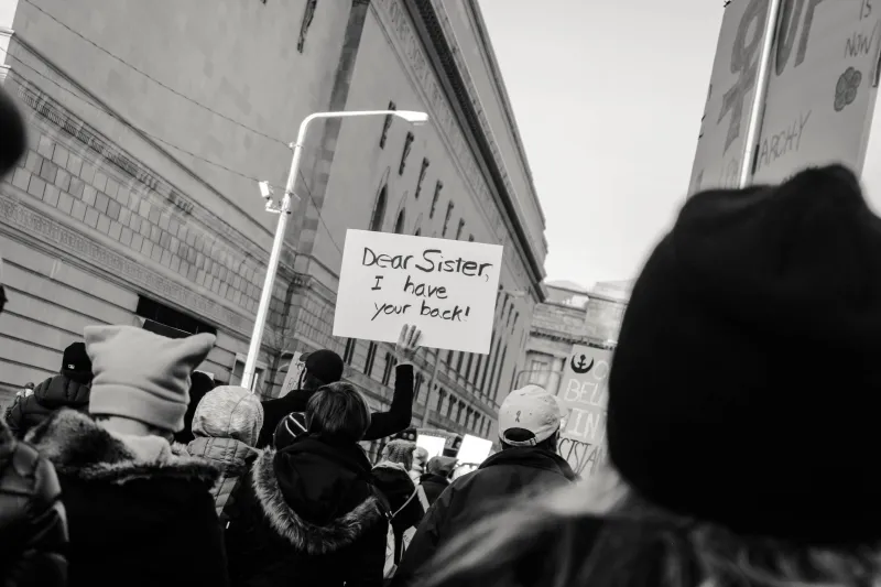 Group of people protesting on street in winter. One sign says, “Dear Sister, I have your back.!”