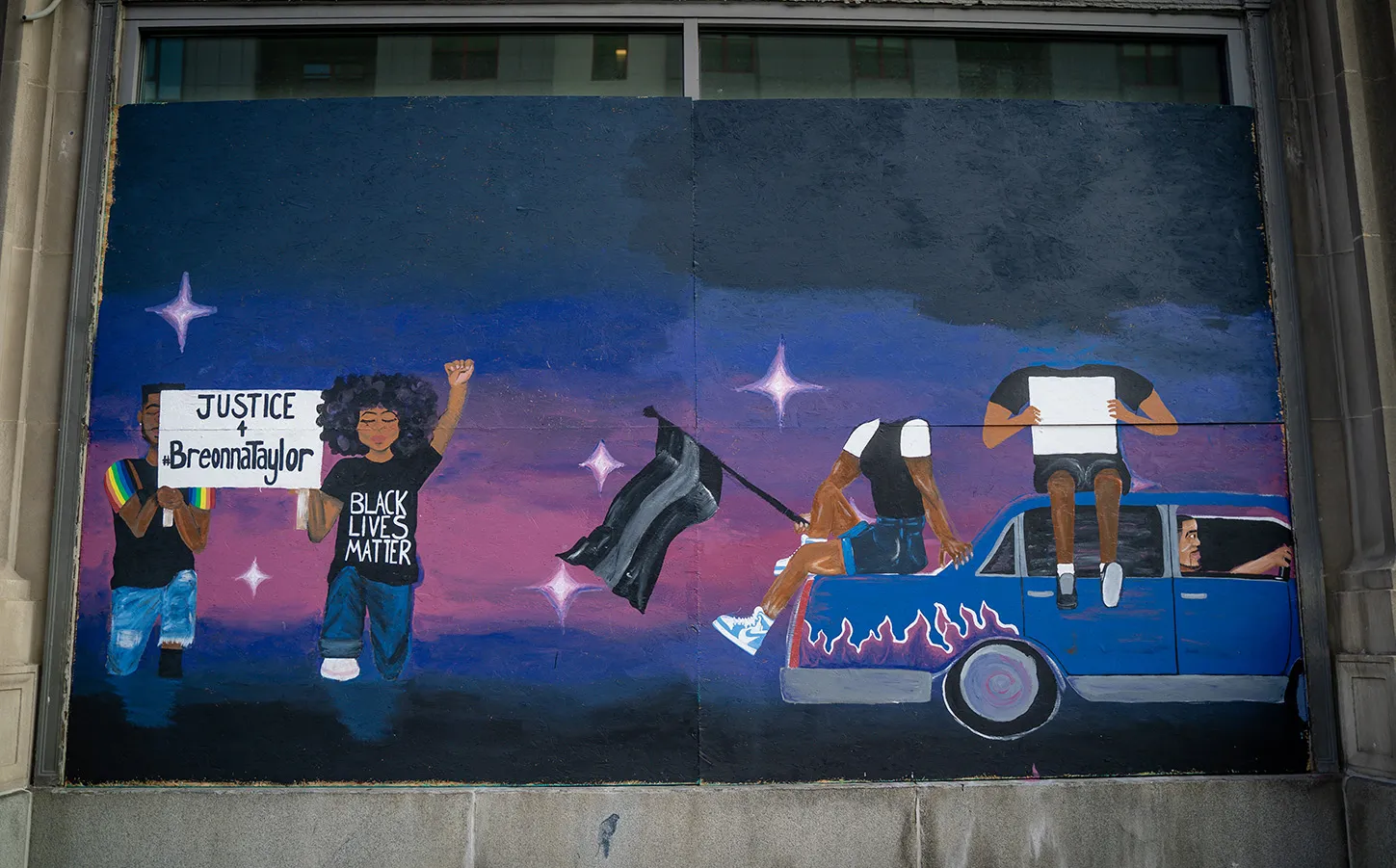 A color photo that is taken front-on. The picture is of street art painted on plywood covering windows on a building downtown Columbus. The background of the art is a top-to-bottom gradient: black, blue, pink, purple, blue, black. The bottom right corner shows two figures holding a sign that reads “Justice for Breonna Taylor.” The bottom right corner shows a blue car with pink and purple flames. Two headless figures sit on the car, third figure is driving the car in the rightward direction. 