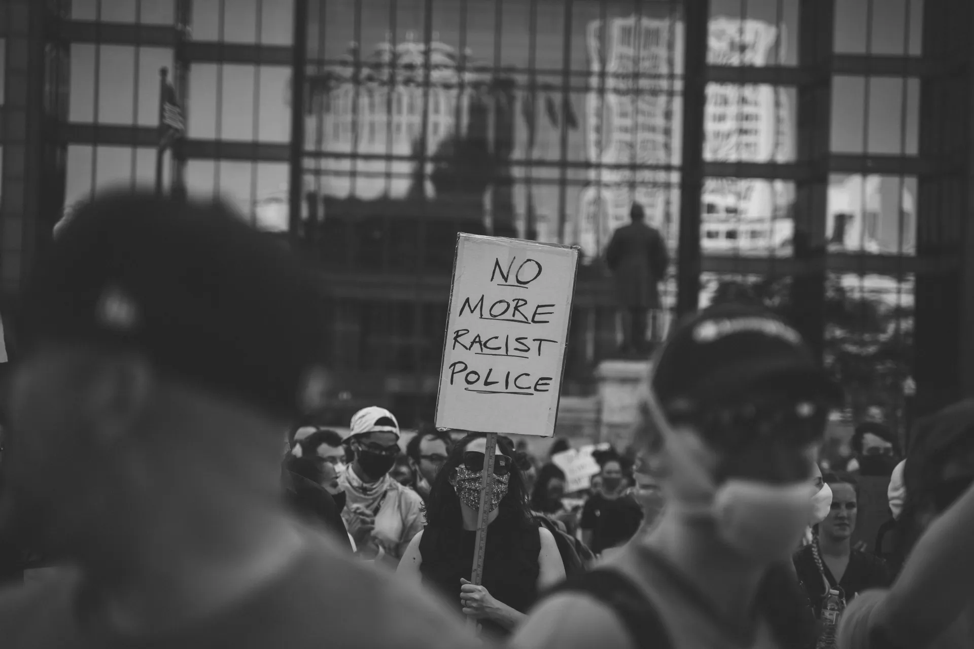 A black and white photo. The image focuses on the middle ground featuring a protester carrying a sign that says "no more racist police." The foreground shows more protestors out of focus. The background shows a building with windows that reflect images of many buildings in downtown Columbus.